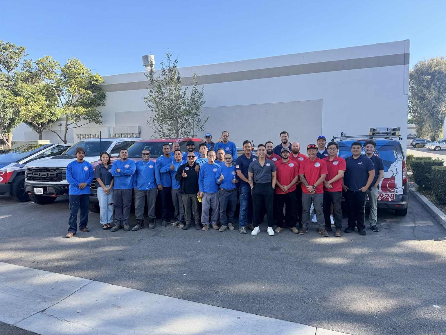 About twenty-five Real Time Bros team members in blue, red and navy shirts, standing in front of their trucks
